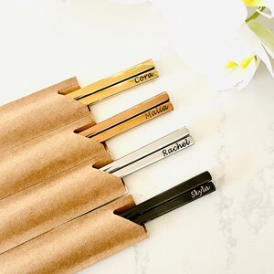 May include: Set of four personalized chopsticks in a brown paper sleeve. The chopsticks are gold, rose gold, silver, and black. The names Cora, Malia, Rachel, and Skyla are engraved on the chopsticks.