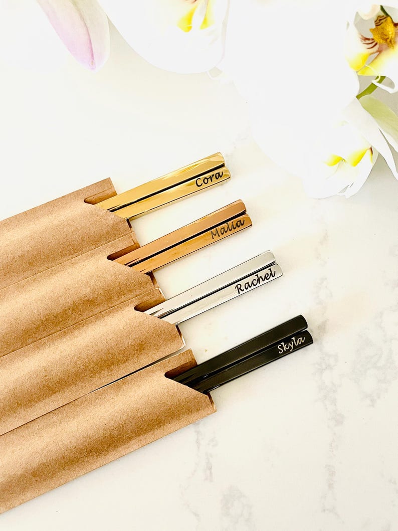 May include: Set of four personalized chopsticks in a brown paper sleeve. The chopsticks are gold, rose gold, silver, and black. The names Cora, Malia, Rachel, and Skyla are engraved on the chopsticks.