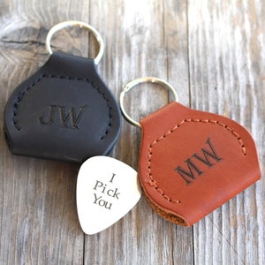 May include: Two leather guitar pick holders with key rings. One is black with the initials "JW" and the other is brown with the initials "MW". A silver guitar pick with the text "I Pick You" is in the center.