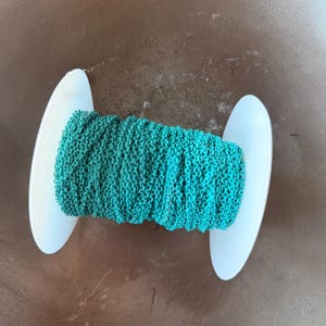 以下が含まれることがあります： ティール色のチェーンの巻き取り。チェーンは白いプラスチック製のスプールにしっかりと巻き付けられています。チェーンは質感があり、色は鮮やかなティールです。スプールは画像の中央に配置されています。