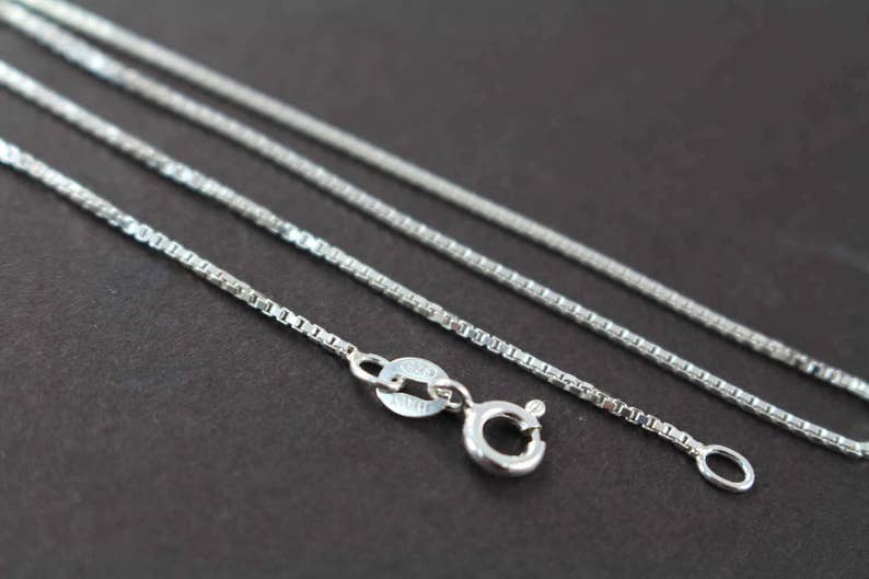 May include: Three silver chains of varying lengths, all with a box chain design. The chains are laid out on a dark surface, with the clasp of the shortest chain visible in the foreground.