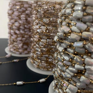 May include: Three spools of beaded chain in shades of pink, gold, and gray. The chain features rectangular beads strung on gold-colored wire. The spools are on white plastic bases, ideal for jewelry making or craft projects.