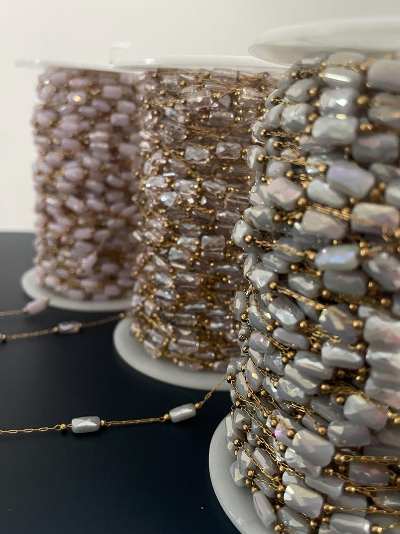May include: Three spools of beaded chain in shades of pink, gold, and gray. The chain features rectangular beads strung on gold-colored wire. The spools are on white plastic bases, ideal for jewelry making or craft projects.