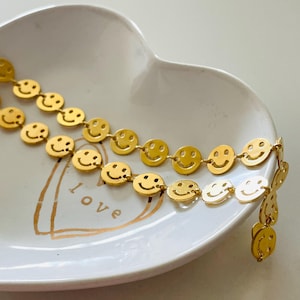 May include: Two gold chains with a repeating pattern of smiley faces. The chains are on a white heart-shaped dish with the word "love" written on it.