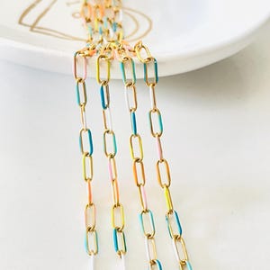 Op de afbeelding: Vijf strengen gouden ketting met kleurrijke emaille accenten. De kettingen zijn verticaal gerangschikt en bevinden zich op een wit oppervlak. De emaille kleuren zijn turquoise, geel, roze en wit.