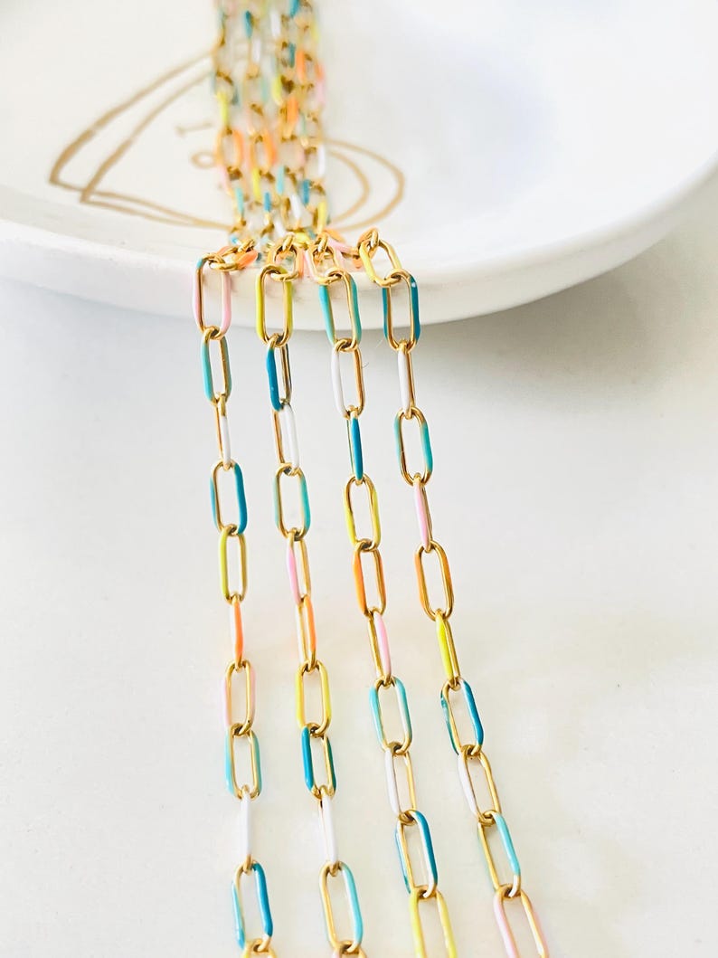 Op de afbeelding: Vijf strengen gouden ketting met kleurrijke emaille accenten. De kettingen zijn verticaal gerangschikt en bevinden zich op een wit oppervlak. De emaille kleuren zijn turquoise, geel, roze en wit.