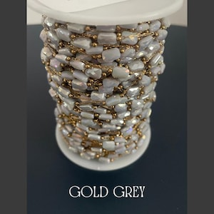 May include: A spool of gold and gray beaded chain. The chain is made of small, rectangular, iridescent gray beads strung together with gold-colored chain. The spool is white, and the text "GOLD GREY" is at the bottom.