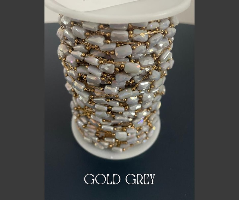 May include: A spool of gold and gray beaded chain. The chain is made of small, rectangular, iridescent gray beads strung together with gold-colored chain. The spool is white, and the text "GOLD GREY" is at the bottom.