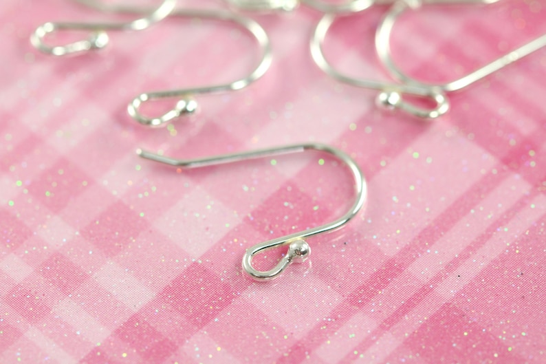 May include: Silver earring hooks with a small ball at the end. The hooks are on a pink glittery surface.