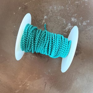 以下が含まれることがあります： ターコイズ色のチェーンが巻かれたスプールで、両端に白いプラスチック製のフランジが付いています。チェーンは鮮やかなティール色で、スプールは茶色の表面に置かれています。