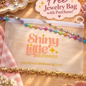Puede incluir: Una bolsa rectangular de pl&aacute;stico transparente con cremallera blanca, que presenta el texto Shiny little BLESSINGS y una URL. Una cadena dorada con dijes de tr&eacute;bol de colores y estrellas descansa encima, con una cadena de pinchos dorada debajo.