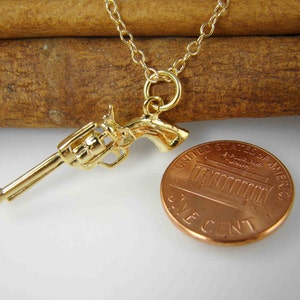 Pode incluir: Um colar com pingente de rev&oacute;lver dourado. O detalhado pingente de arma est&aacute; suspenso em uma delicada corrente dourada. Uma moeda americana est&aacute; ao lado do pingente para compara&ccedil;&atilde;o de tamanho. Fundo claro.