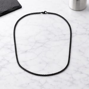 May include: A black chain necklace with a box link design. The necklace is made of a dark metal and is displayed on a white and grey marble surface. The necklace is a simple, elegant piece of jewellery.
