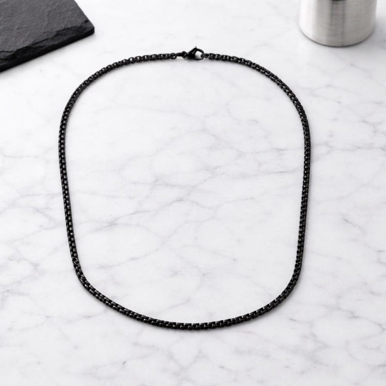 May include: A black chain necklace with a box link design. The necklace is made of a dark metal and is displayed on a white and grey marble surface. The necklace is a simple, elegant piece of jewellery.