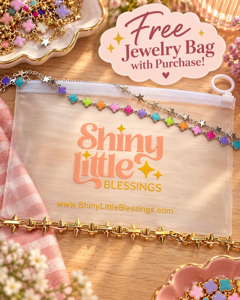 Puede incluir: Una bolsa rectangular de pl&aacute;stico transparente con cremallera blanca, que presenta el texto Shiny little BLESSINGS y una URL. Una cadena dorada con dijes de tr&eacute;bol de colores y estrellas descansa encima, con una cadena de pinchos dorada debajo.