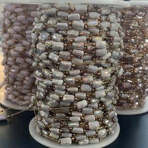 May include: Spools of beaded chain in various colors. The primary spool features a light gray chain with rectangular beads and gold-colored chain links. Other spools display similar designs in different shades, including a pale purple and a gold-toned chain.