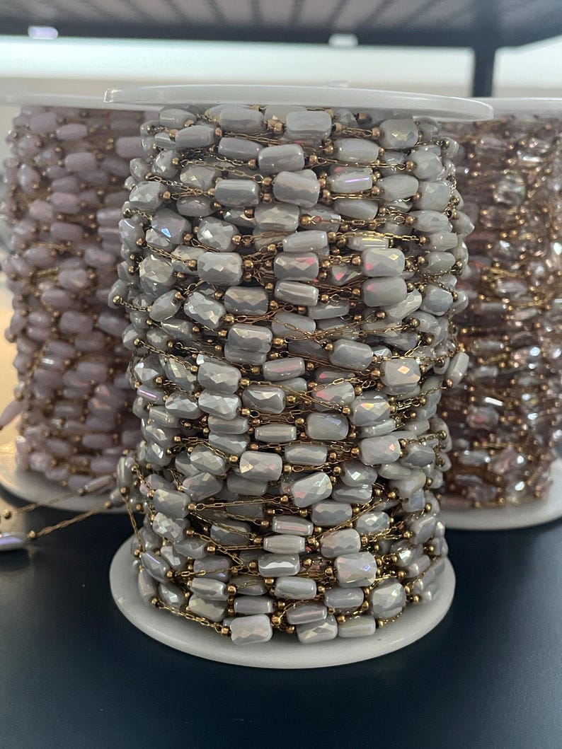 May include: Spools of beaded chain in various colors. The primary spool features a light gray chain with rectangular beads and gold-colored chain links. Other spools display similar designs in different shades, including a pale purple and a gold-toned chain.