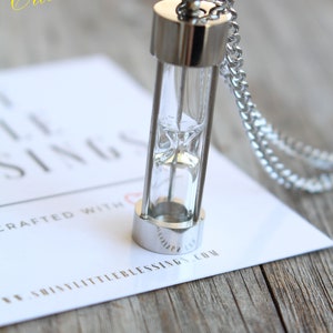 May include: A silver hourglass necklace with a clear glass vial and a silver chain. The necklace is on a white card with the text "Custom Engraved" and "Shiny Little Blessings.com".