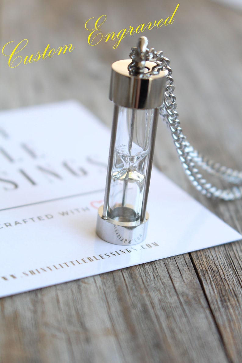 May include: A silver hourglass necklace with a clear glass vial and a silver chain. The necklace is on a white card with the text "Custom Engraved" and "Shiny Little Blessings.com".