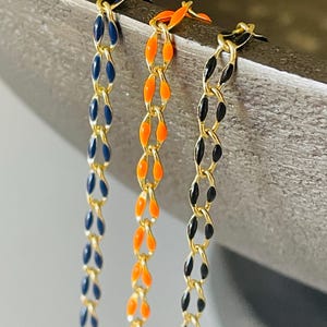 May include: Three gold-toned chain necklaces with colorful enamel links. One is navy blue, one is orange, and the third is black. The chains are hanging against a neutral background.