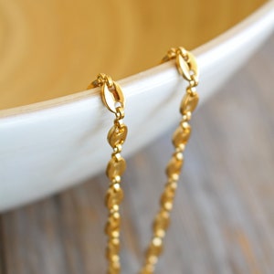 May include: A gold chain necklace with a delicate, oval-shaped link design. The chain is draped over the edge of a white bowl.