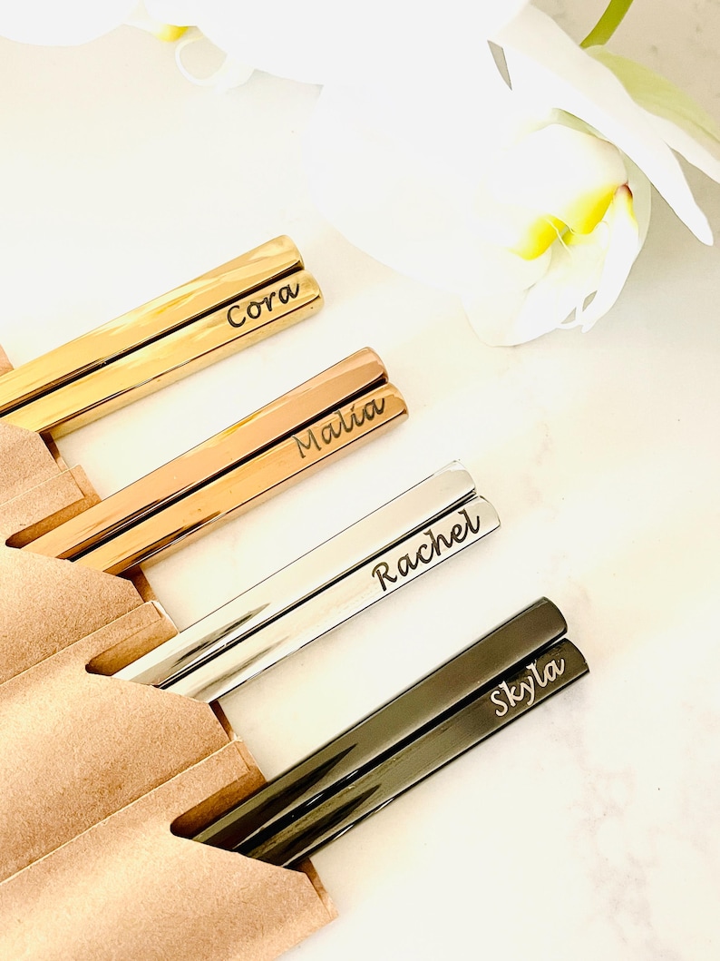 May include: Four pairs of personalized chopsticks in gold, rose gold, silver, and black. The chopsticks are engraved with the names Cora, Malia, Rachel, and Skyla.