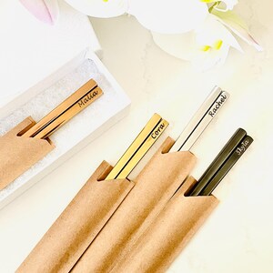 May include: Four pairs of personalized chopsticks in brown paper sleeves. The chopsticks are gold, silver, and black. The names "Malia", "Cora", "Rachel", and "Skyla" are engraved on the chopsticks.