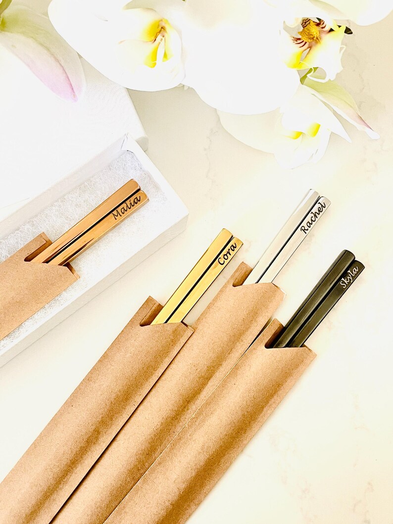 May include: Four pairs of personalized chopsticks in brown paper sleeves. The chopsticks are gold, silver, and black. The names "Malia", "Cora", "Rachel", and "Skyla" are engraved on the chopsticks.