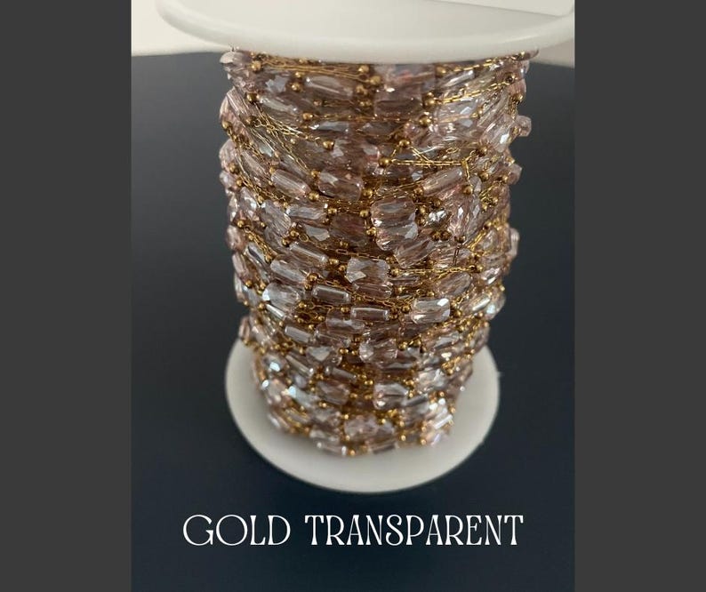 May include: A spool of gold-colored chain with transparent, rectangular beads. The chain is wound around a white spool. The text "GOLD TRANSPARENT" is at the bottom of the image.