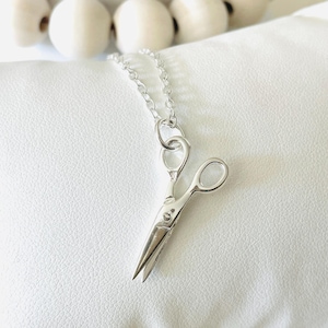 May include: A silver necklace with a small, detailed silver scissor charm. The necklace is on a white background.