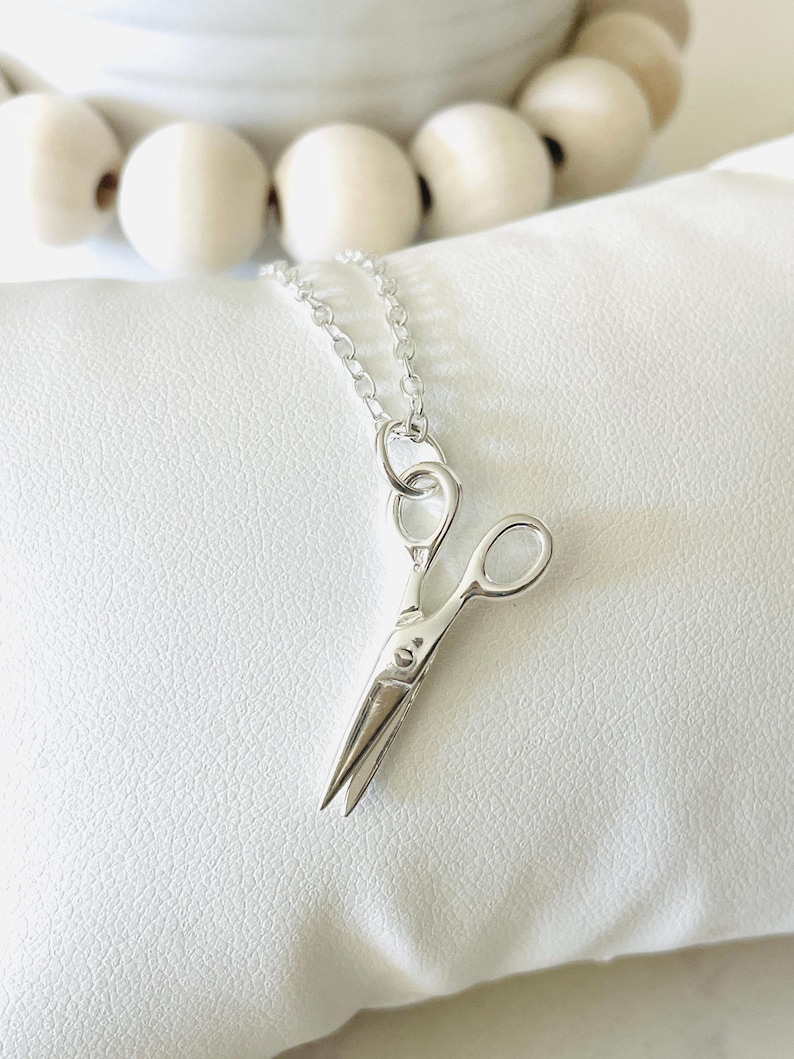 May include: A silver necklace with a small, detailed silver scissor charm. The necklace is on a white background.