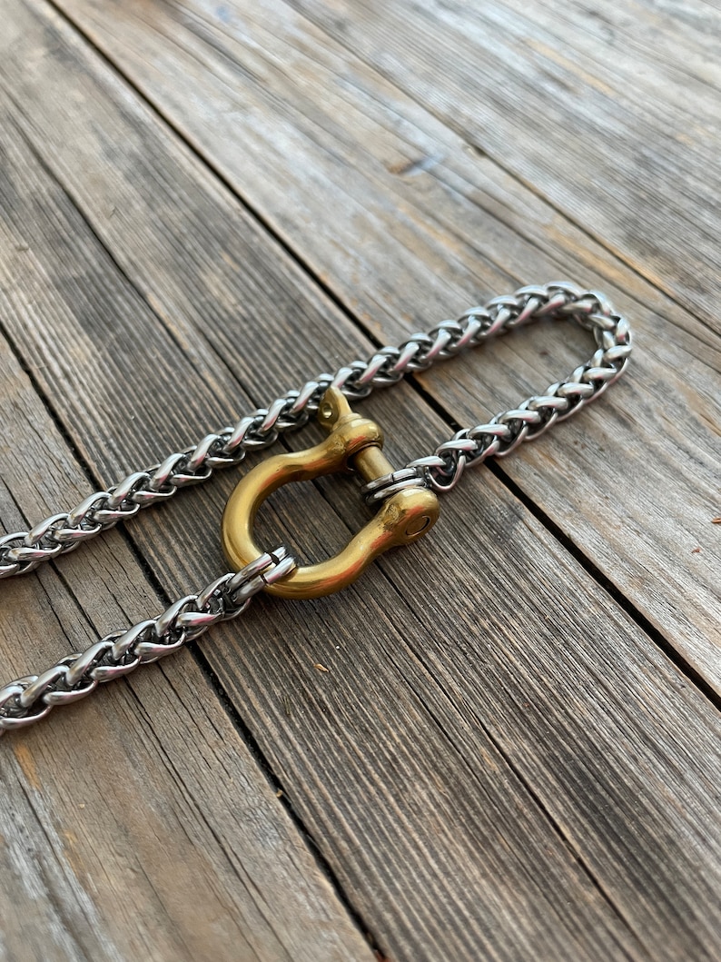 Pu&ograve; includere: Una catena d'argento con una chiusura a forma di U dorata. La catena e la chiusura sono su una superficie di legno.