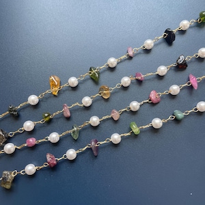 Puede incluir: Primer plano de varias hebras de joyer&iacute;a con una cadena dorada, perlas blancas y piedras preciosas de colores de forma irregular. Las piedras preciosas son en tonos rosa, verde, amarillo y negro. El fondo es azul oscuro liso.