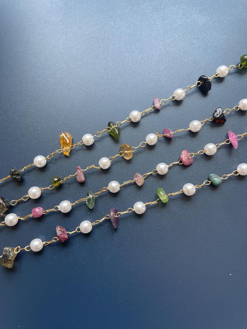 Puede incluir: Primer plano de varias hebras de joyer&iacute;a con una cadena dorada, perlas blancas y piedras preciosas de colores de forma irregular. Las piedras preciosas son en tonos rosa, verde, amarillo y negro. El fondo es azul oscuro liso.