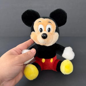 May include: A plush Mickey Mouse toy with black fur, large black ears, and a tan face. The toy has a black body, red shorts with yellow buttons, and yellow feet. The toy is being held by a hand.