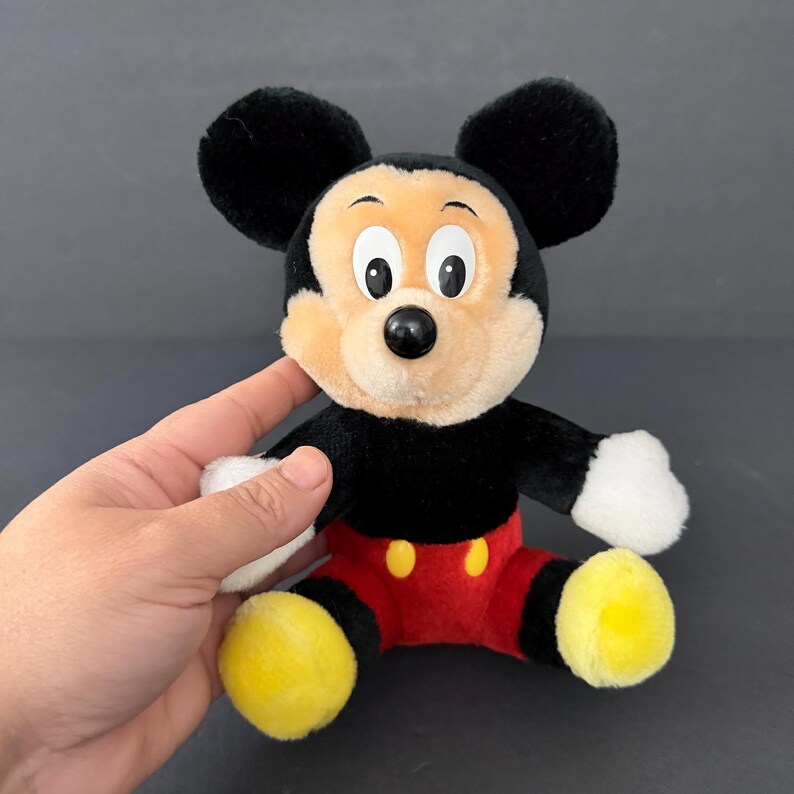 May include: A plush Mickey Mouse toy with black fur, large black ears, and a tan face. The toy has a black body, red shorts with yellow buttons, and yellow feet. The toy is being held by a hand.