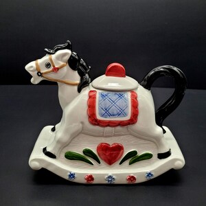 Peut inclure: Théière en céramique blanche en forme de cheval à bascule, avec crinière et queue noires. La théière présente une selle rouge et bleue, un cœur rouge et des motifs floraux. Objet décoratif.