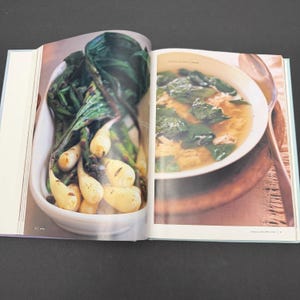 May include: Open cookbook pages with a white bowl of roasted vegetables and a bowl of soup. The vegetables are white and green, and the soup has green leaves. The cookbook is open on a black surface.