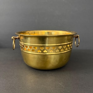 May include: A brass planter with a heart-shaped cutout design around the middle. The planter has two ring handles and a shiny, golden finish. The planter is a decorative item.