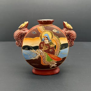 May include: A decorative, round, ceramic vase with a red base and handles. The vase features a detailed illustration of a figure with a dragon, set against a landscape with blue water and an orange sky. The vase is adorned with gold accents and white polka dots.