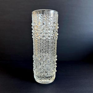 Könnte beinhalten: Eine transparente Glasvase mit strukturierter Oberfläche. Die Vase hat eine zylindrische Form und ist mit kleinen, abgerundeten Noppen bedeckt. Das Glas ist transparent, so dass Licht hindurchscheinen kann. Geeignet für Blumen oder als dekoratives Element.