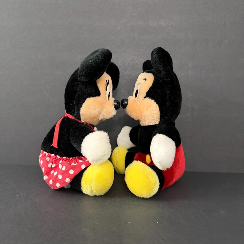 May include: Two plush Disney characters, Mickey and Minnie Mouse, face each other. Mickey has a red and yellow outfit, while Minnie wears a red polka-dot skirt. Both have black fur, tan faces, and black ears.
