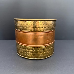 May include: A cylindrical container with a brass top and bottom, and a copper band in the middle. The brass sections have a textured, dimpled pattern. The container is likely for storage or decoration.