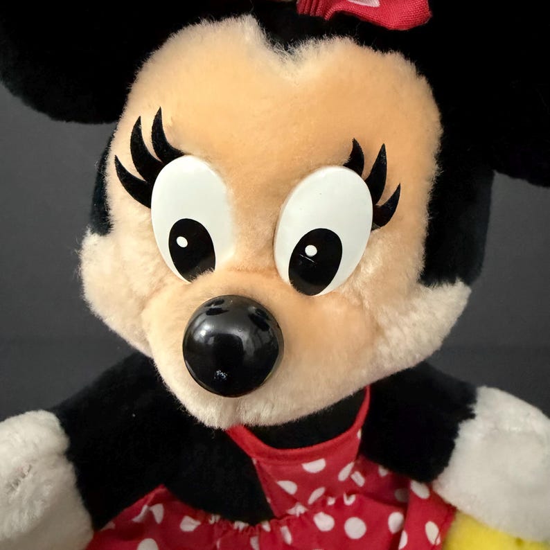 May include: Close-up of a Minnie Mouse plush toy. The plush features a black nose, large white eyes with black pupils, and black eyelashes. She wears a red dress with white polka dots and a red bow on her head. The plush has black ears and a cream-colored face.
