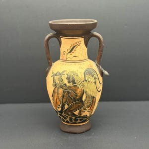 May include: A tan and brown ceramic vase with two handles and a wide mouth. The vase features a black and orange illustration of a figure with wings playing a harp. The word "SELANA" is written on the vase.