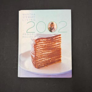 May include: A cookbook titled "Martha Stewart Living 2002 Annual Recipes" with a photo of a layered cake on a white plate. The cover has a light green and blue gradient background with the year 2002 in large numbers and a photo of Martha Stewart.