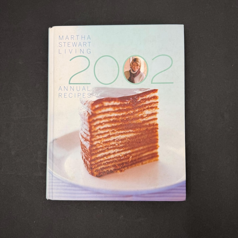 May include: A cookbook titled "Martha Stewart Living 2002 Annual Recipes" with a photo of a layered cake on a white plate. The cover has a light green and blue gradient background with the year 2002 in large numbers and a photo of Martha Stewart.