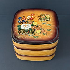 Puede incluir: Una caja bento cuadrada de madera de varios niveles con una tapa decorativa. La tapa presenta un diseño floral con flores blancas, rojas, amarillas y azules, y un borde marrón oscuro. La caja tiene una combinación de colores marrón claro y marrón oscuro.
