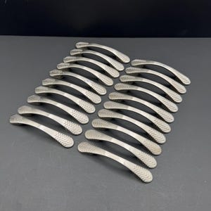 May include: A collection of twenty brushed nickel cabinet pulls arranged in a grid. Each handle features a curved, arched design with a textured, chevron-patterned detail on both ends, providing a consistent look for furniture or cabinetry hardware.