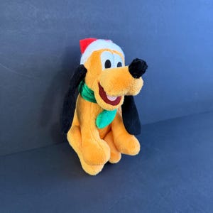 May include: A plush toy of a yellow dog with long black ears and a black nose. The dog is wearing a red and white Santa hat and a green scarf around its neck. The toy has a wide, open-mouthed smile with a pink tongue visible inside the mouth.